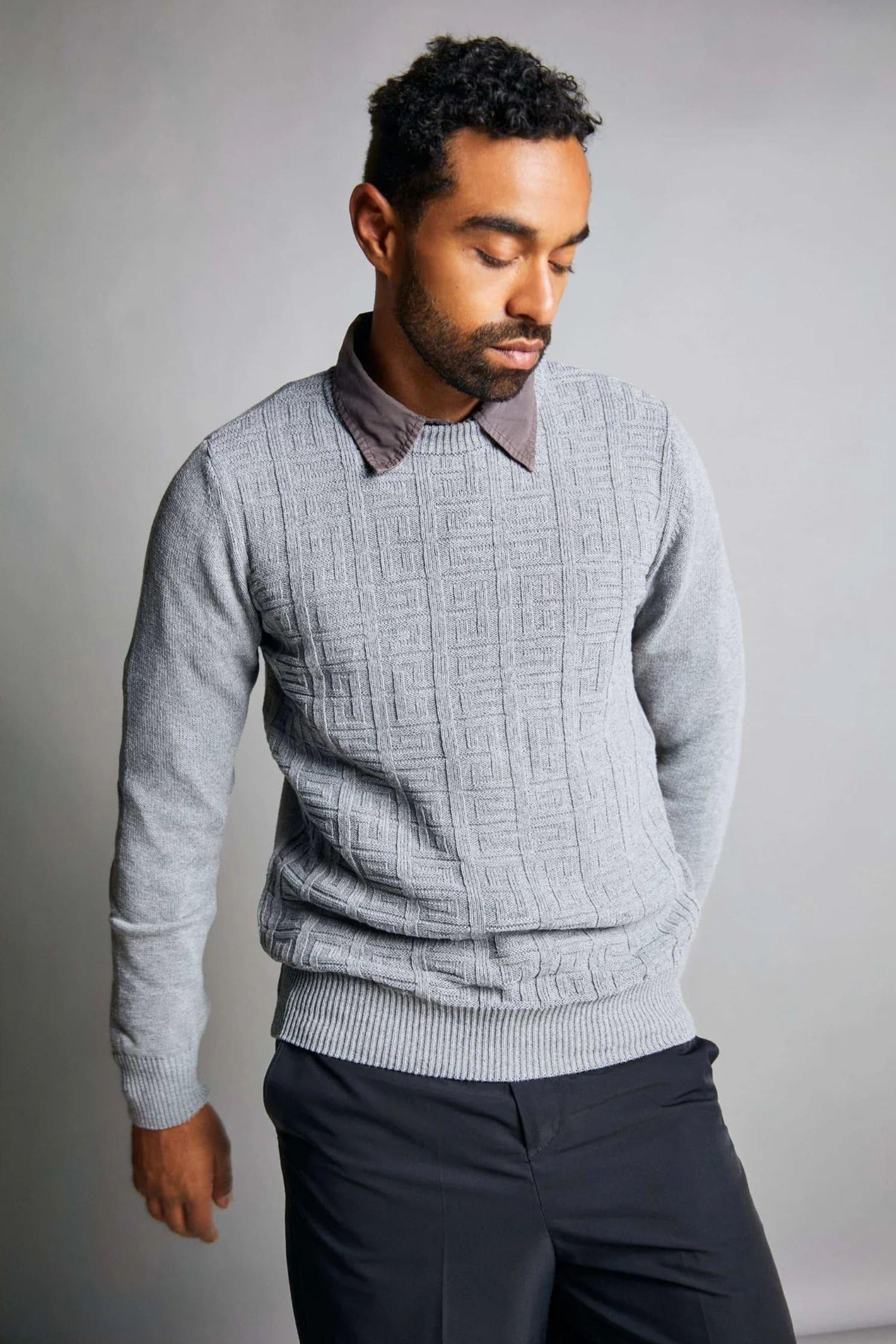 Hombre con suéter gris de punto con diseño geométrico, camisa marrón y pantalón oscuro.